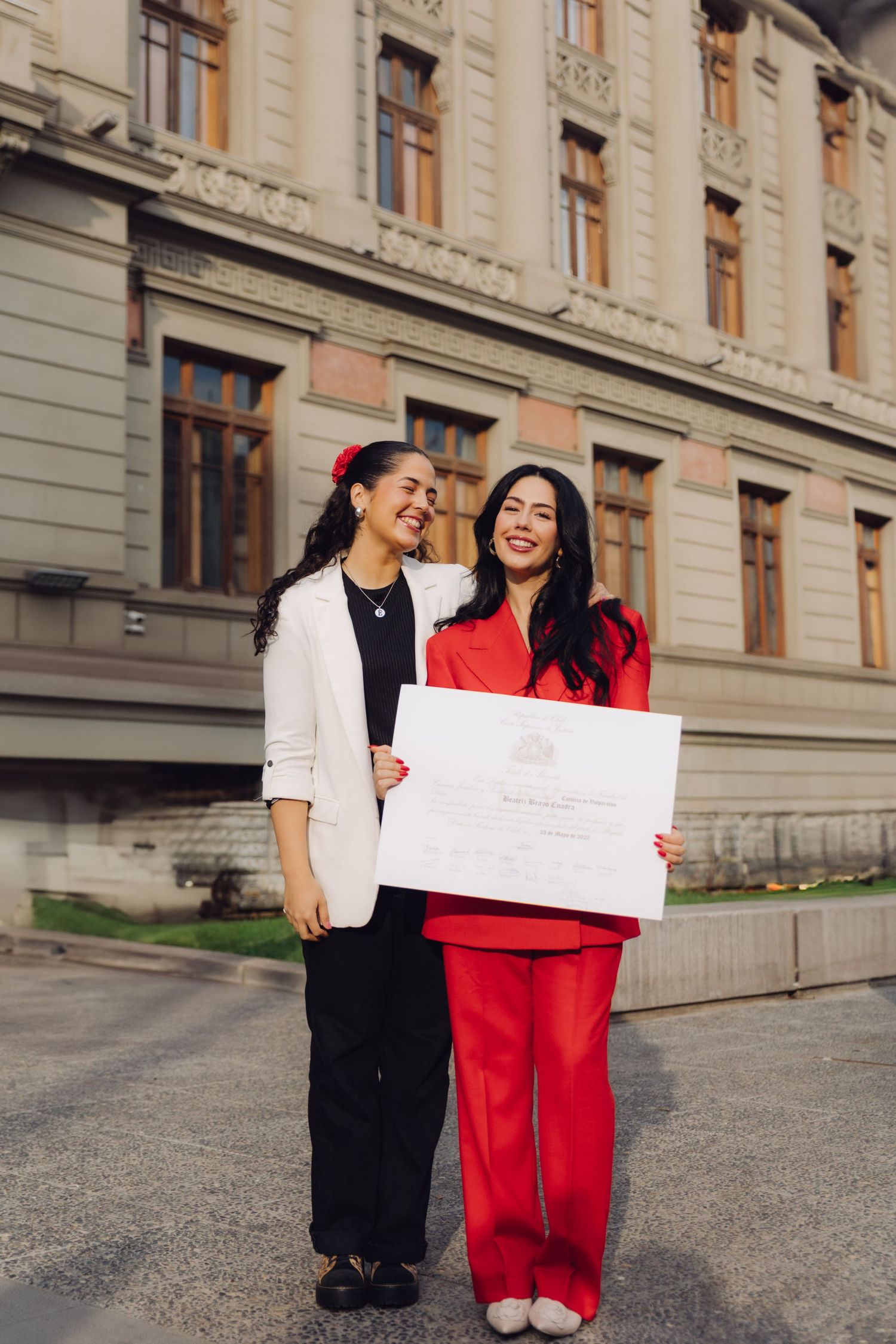 Two women smile proudly outside a historic building, one in red holding a graduation diploma.