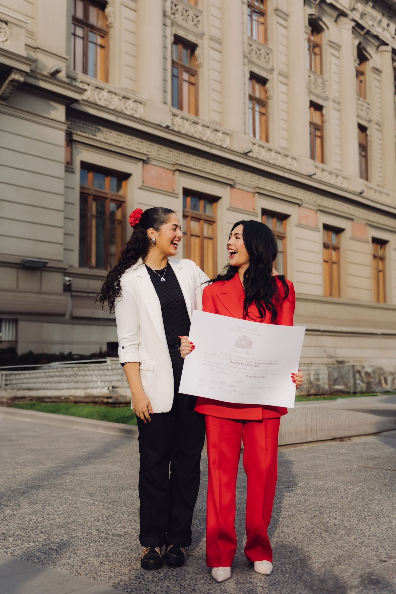 Two laughing women stand outside a classic building, one in red holding a large diploma certificate.
