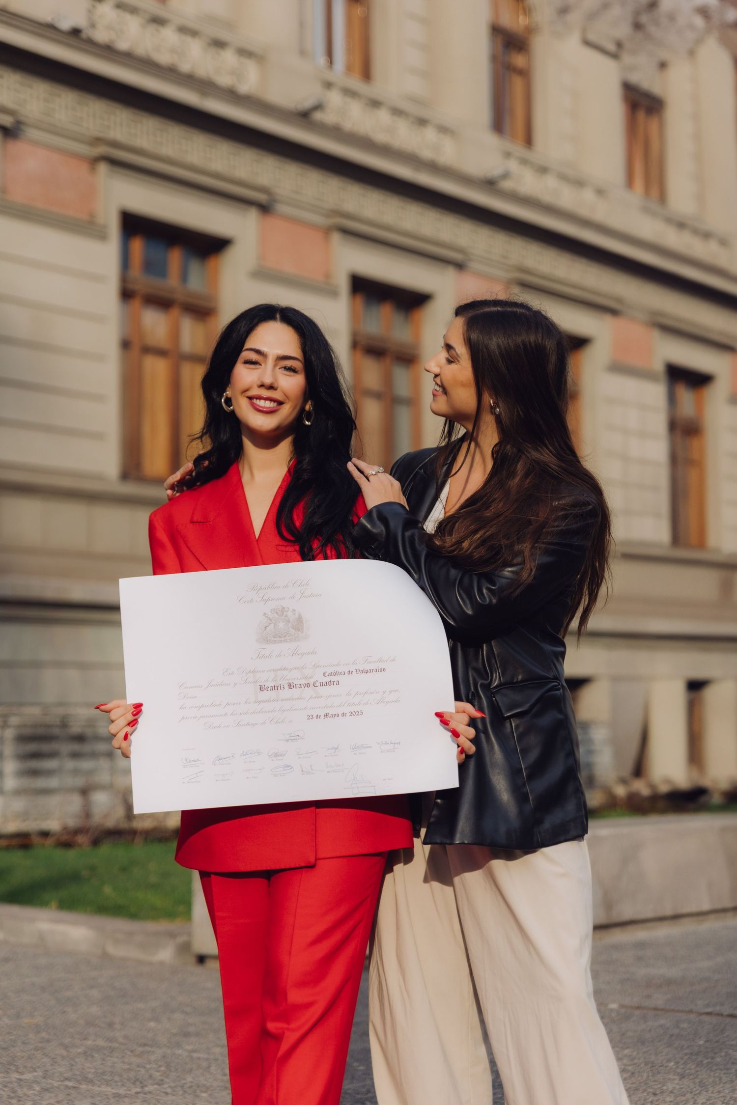 Two women celebrate outdoors holding a diploma, one in red suit and one in black leather jacket.