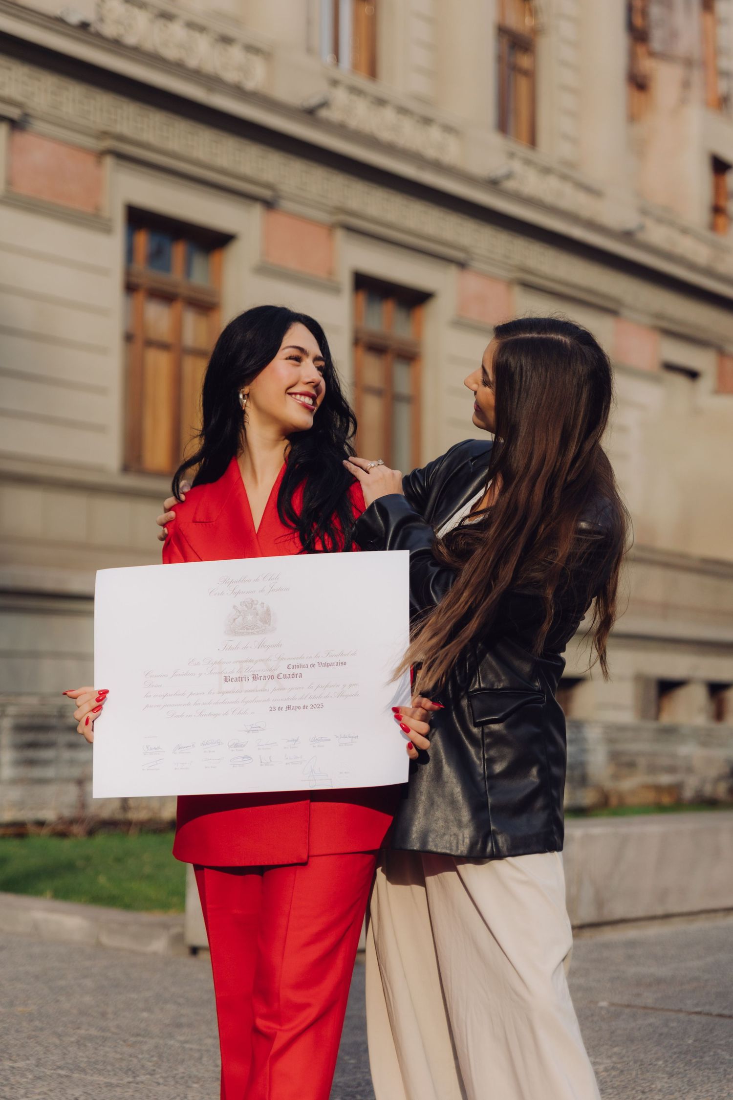 Two women share a joyful moment with a diploma outside a classical stone building.