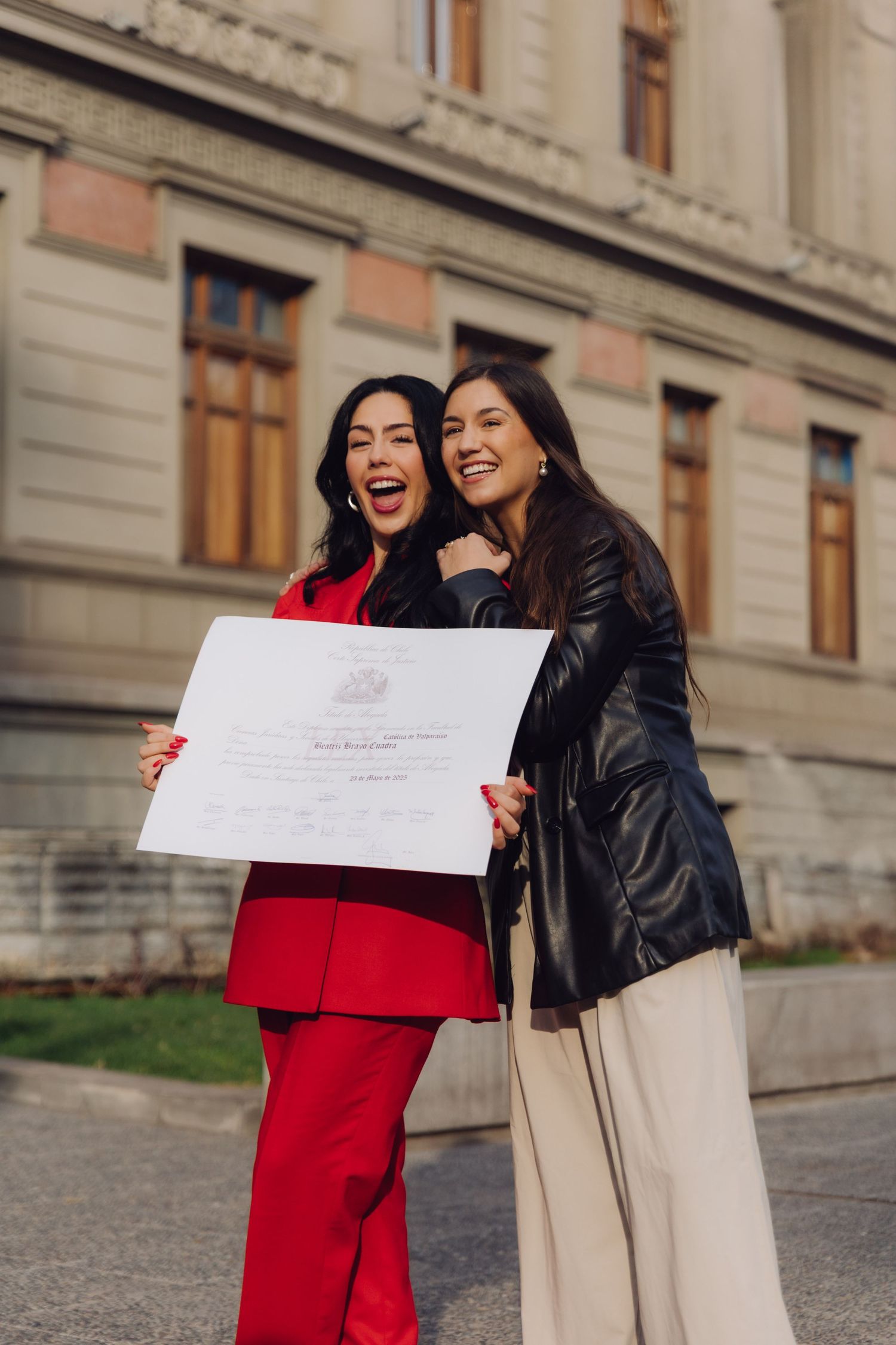 Two women laugh and embrace while holding a graduation certificate outside a historic building.