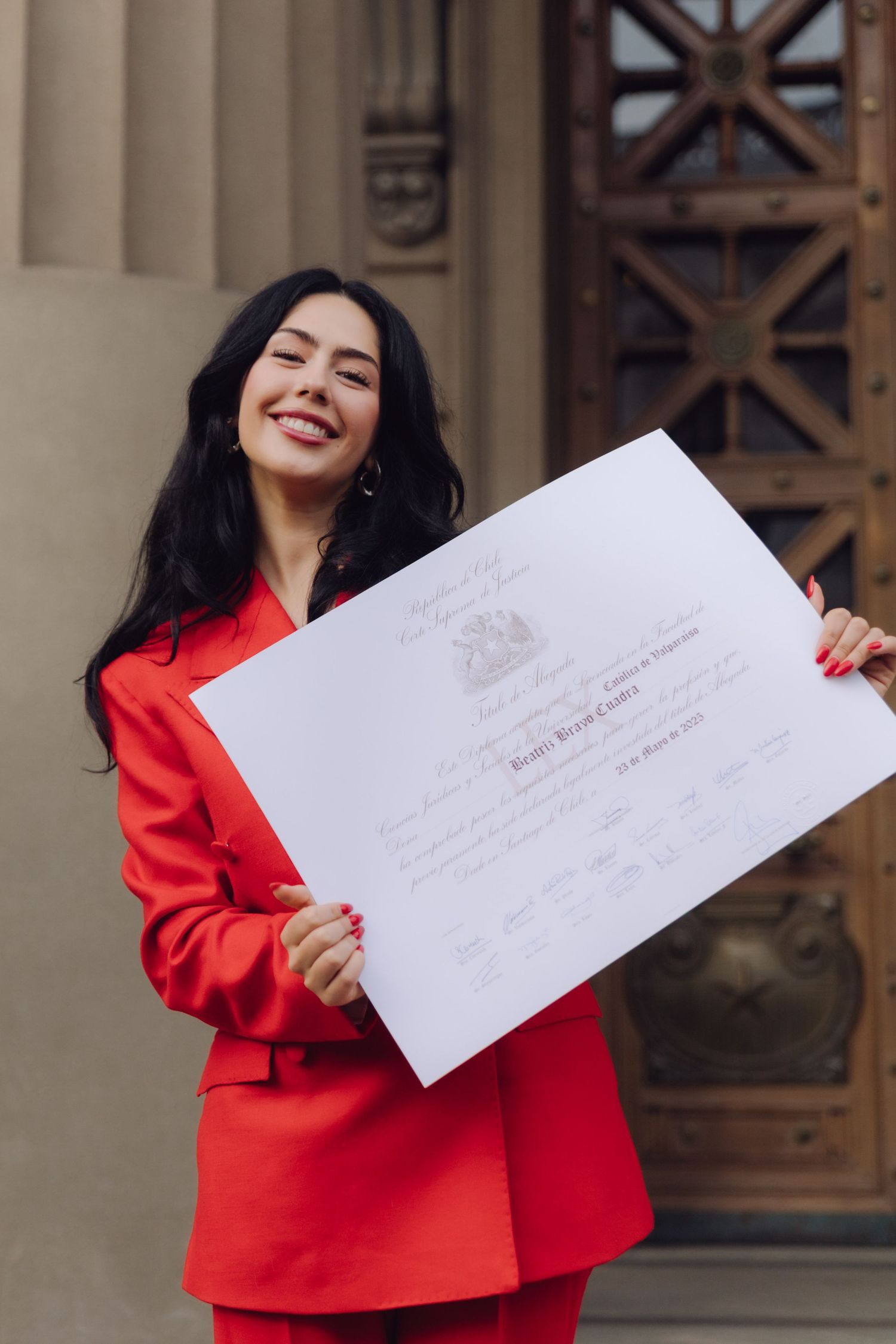Smiling woman in red suit holding a certificate in front of ornate wooden doors.