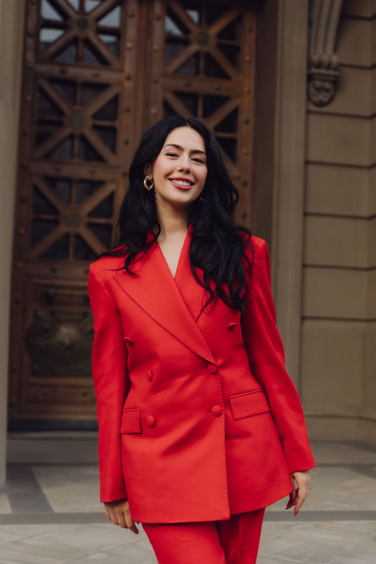 Elegant woman in a bold red suit smiling outside a historic building entrance.