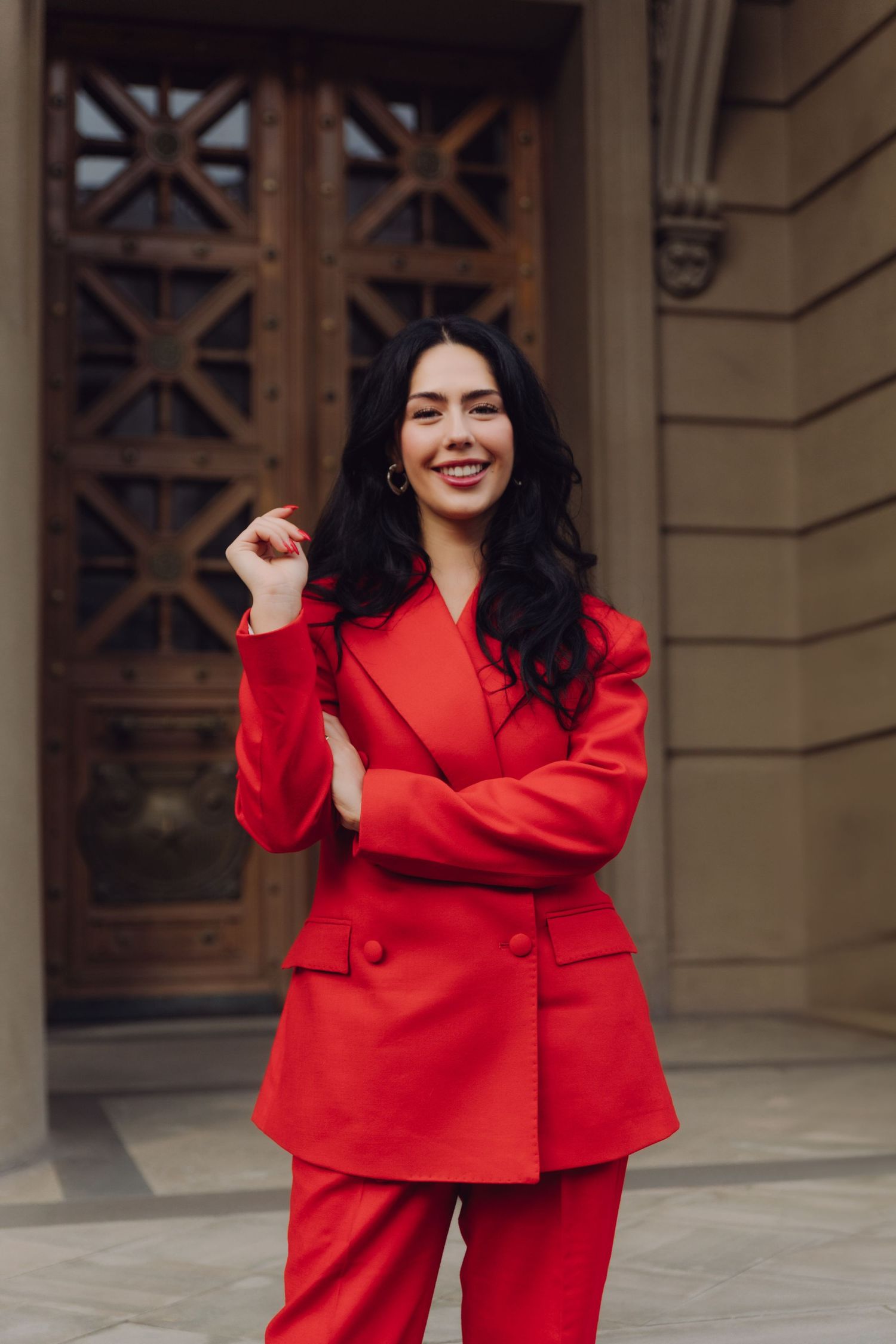 Cheerful woman in red suit posing with arms crossed outside a wooden door.