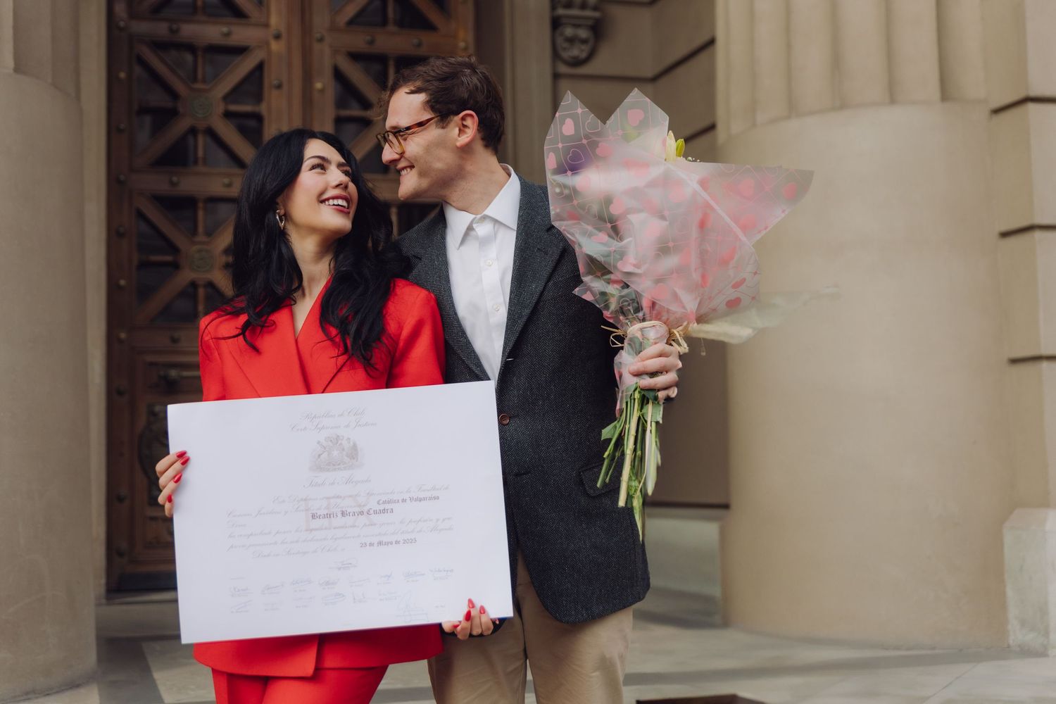 Couple celebrating graduation outdoors, woman in red suit holding diploma certificate, man in suit holding pink flower bouquet.