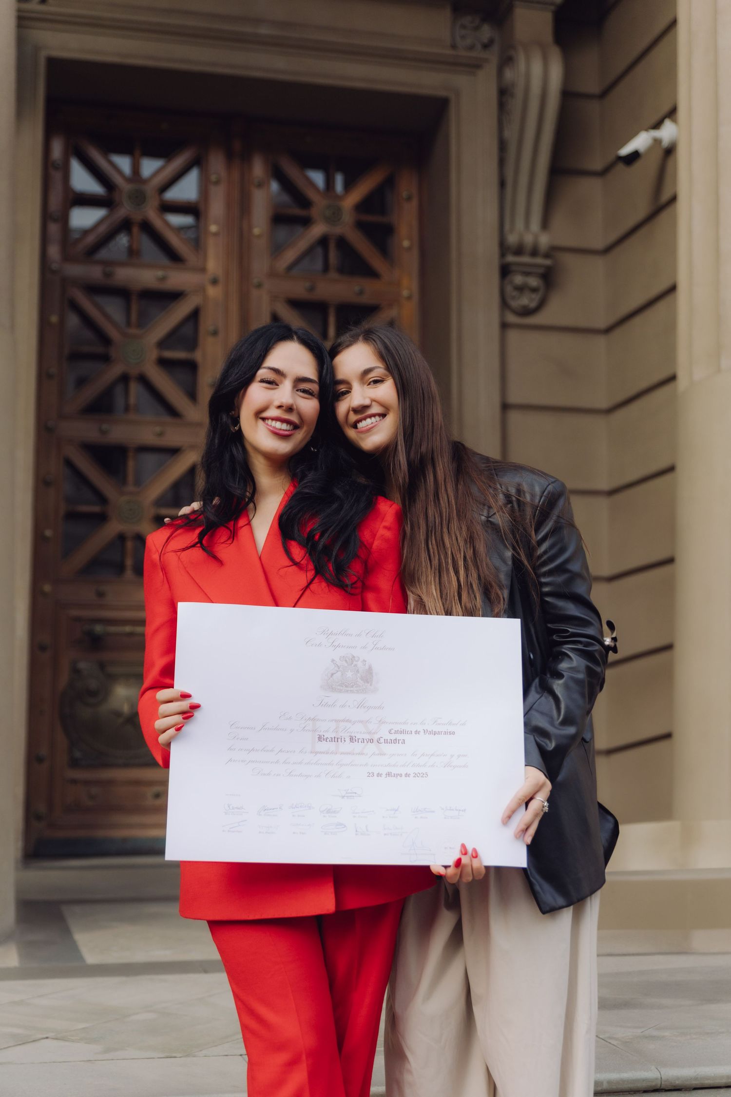 Two women smiling and posing together outdoors holding a large graduation diploma certificate, one wearing a red suit.