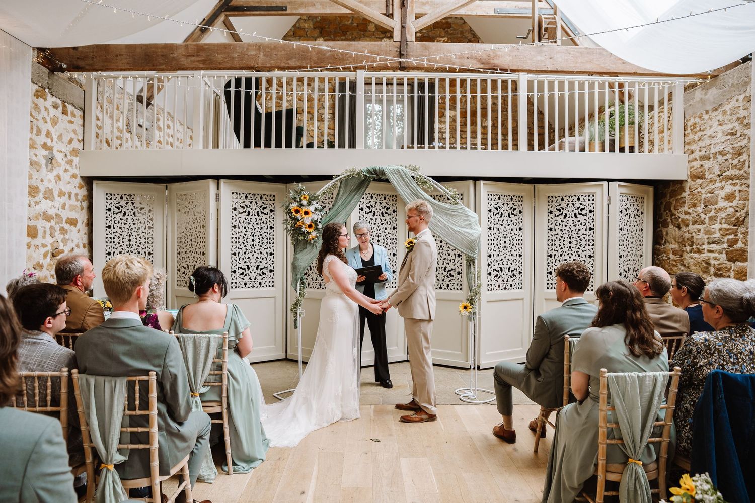 Couple exchanging vows at rustic barn wedding ceremony with stone walls, wooden beams, and seated guests in sage green attire Symmondsbury