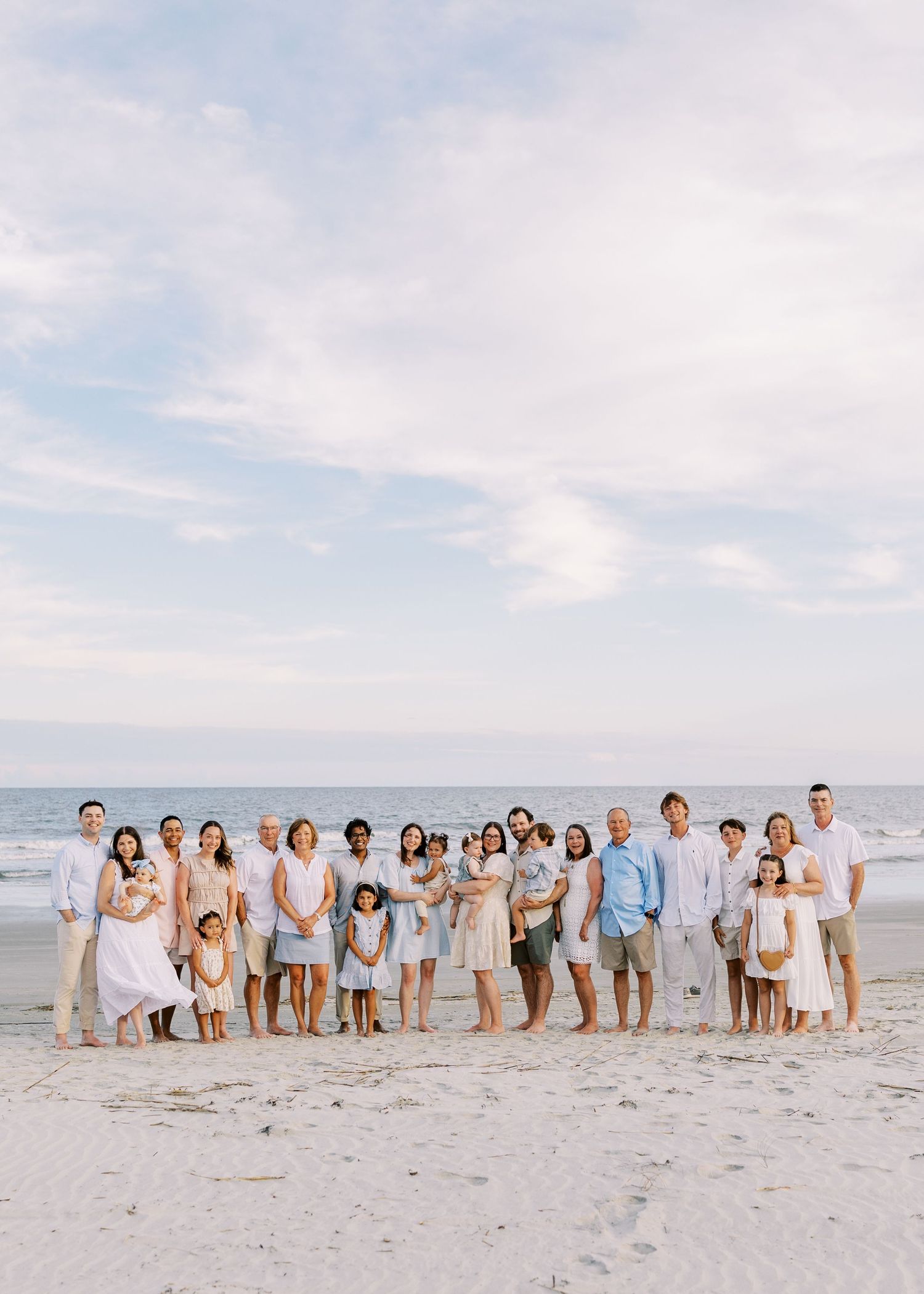 Large family group portrait on a sandy beach at sunset, wearing coordinated white and blue outfits.