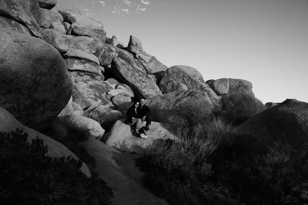 Black and white photo of a person sitting among large boulders in a rocky desert landscape at dusk.