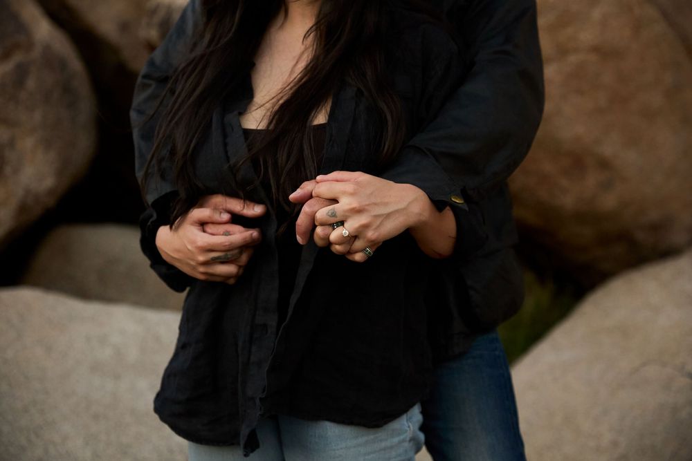 Couple holding hands tenderly, woman wearing gold rings, dressed in black jacket and jeans, with large boulders in background.