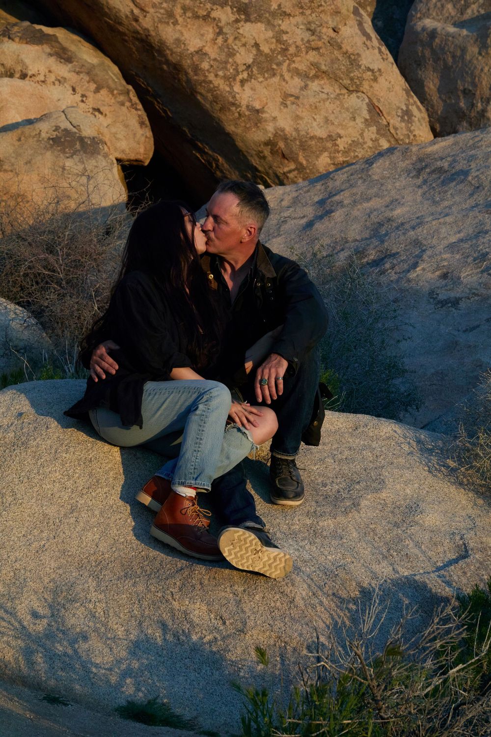 Couple sharing a romantic kiss while sitting on large boulders outdoors at sunset.