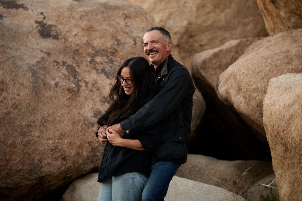 A laughing couple embraces among large desert boulders, both wearing black jackets and jeans outdoors.