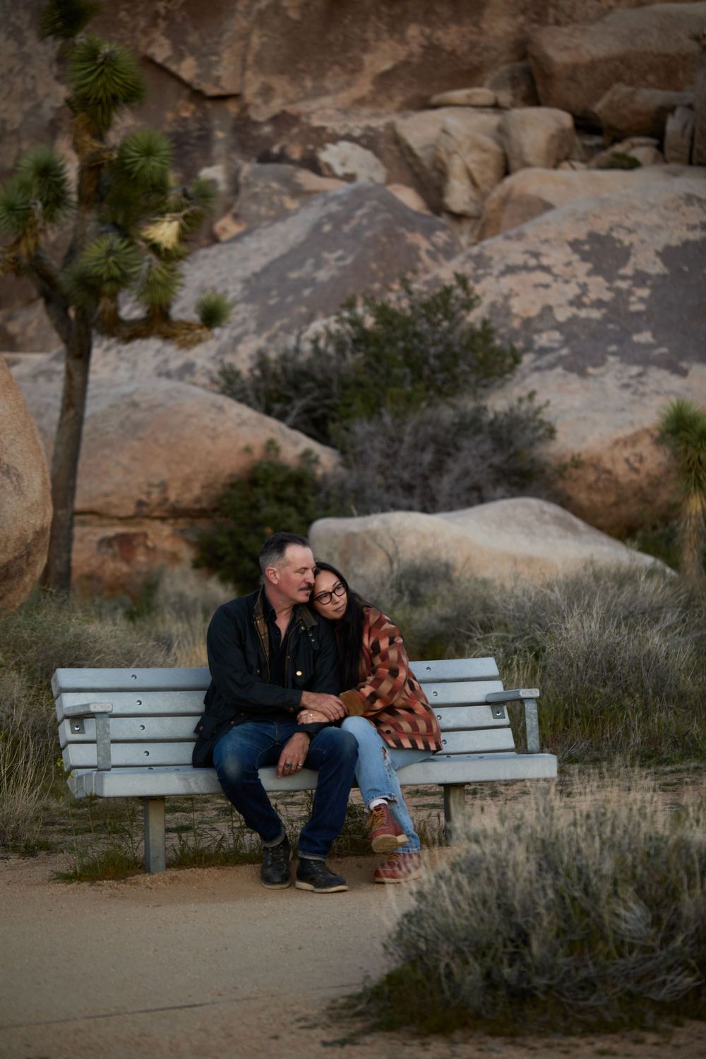 Couple sitting close together on a white bench at Joshua Tree National Park, surrounded by desert rocks and Joshua trees.