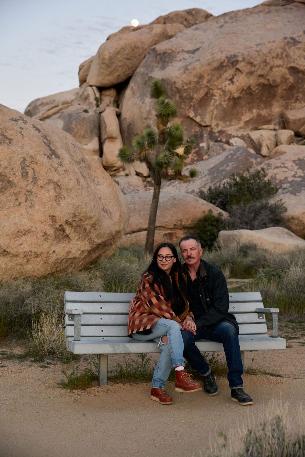 Couple sitting together on a white bench in Joshua Tree National Park with large boulders and a Joshua tree behind them.
