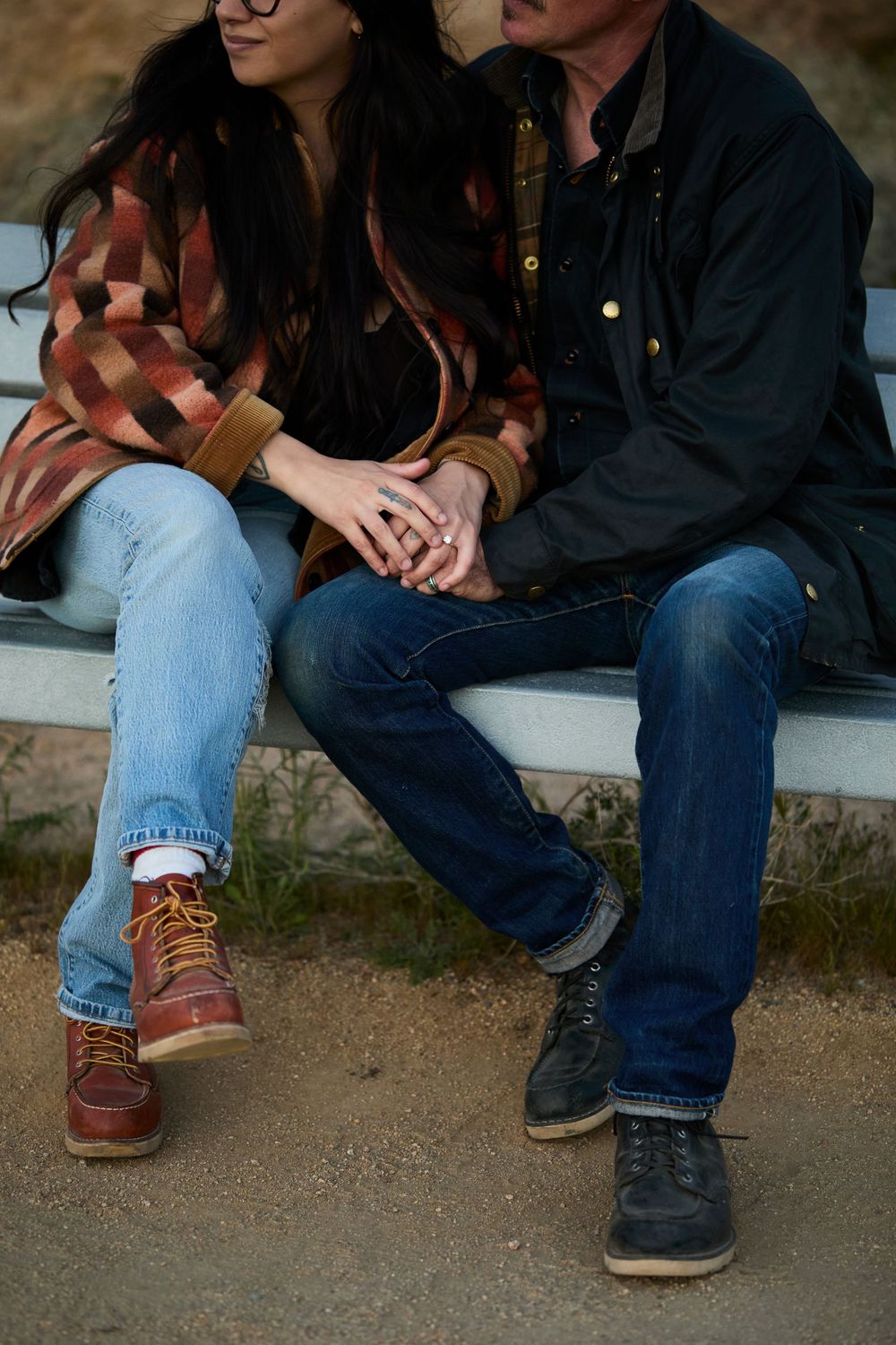 Couple sitting close together on a bench, holding hands, wearing jeans and boots, in a casual outdoor setting.