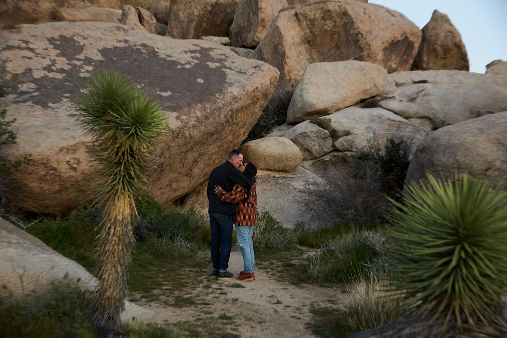 Couple embracing among desert boulders and yucca plants in Joshua Tree National Park at dusk.
