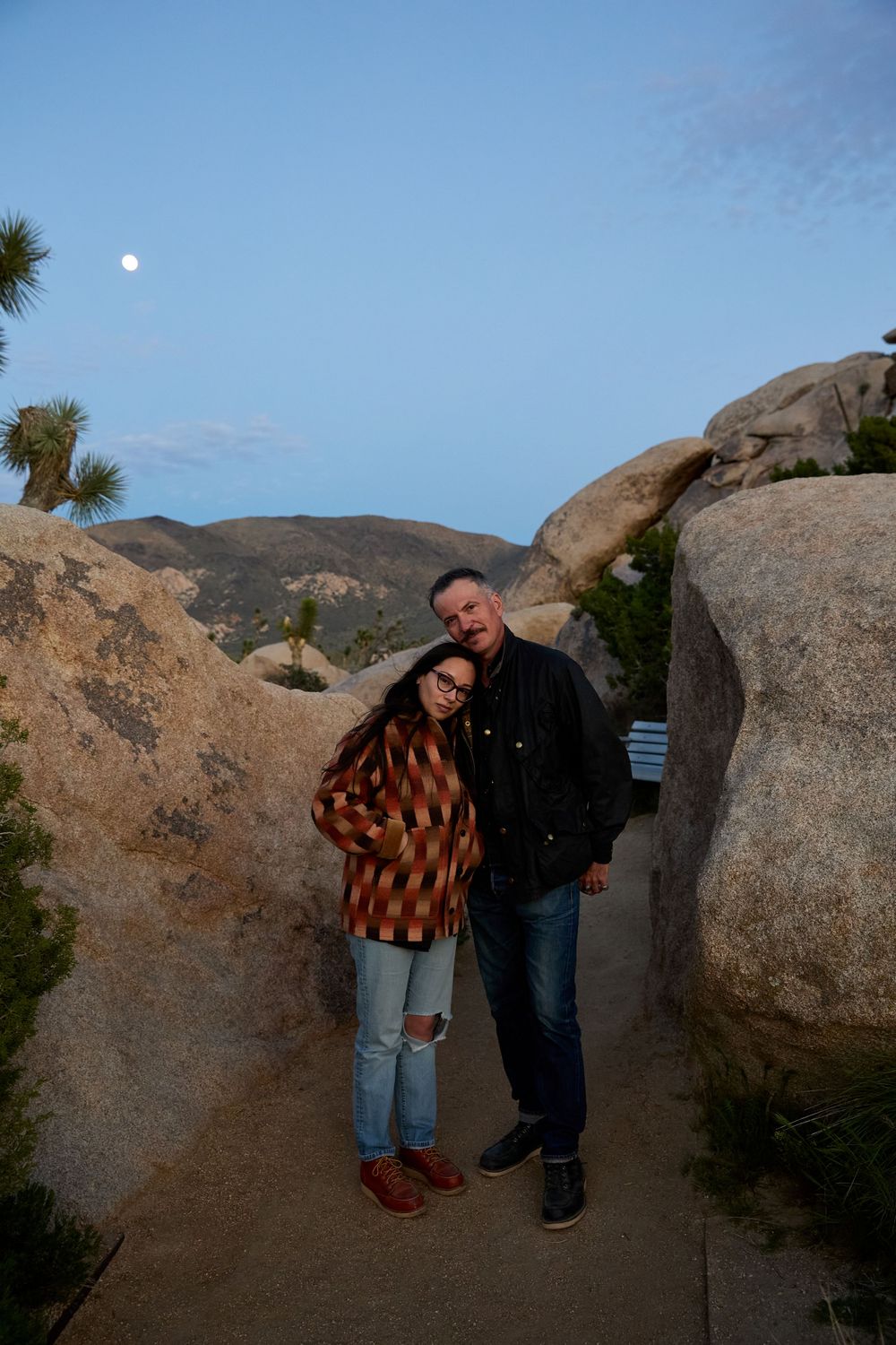 Couple poses between large boulders at dusk in Joshua Tree National Park with moon visible in blue sky.