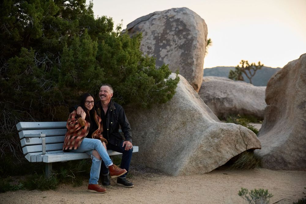 Couple sitting on a bench near large boulders and trees at dusk in a desert landscape.