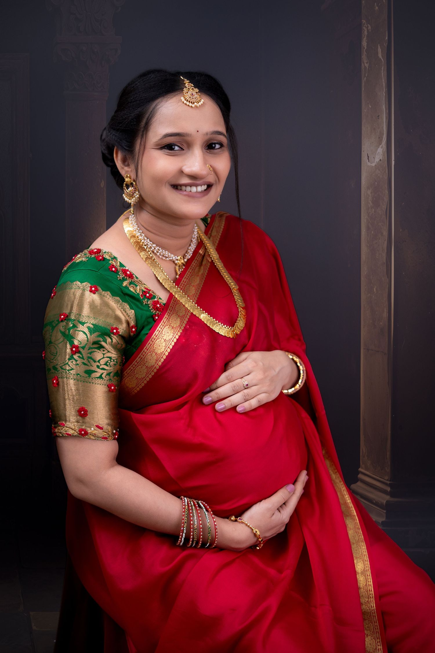 Expecting mother in a red silk saree with a gold-bordered blouse and temple jewellery, hand resting on the bump