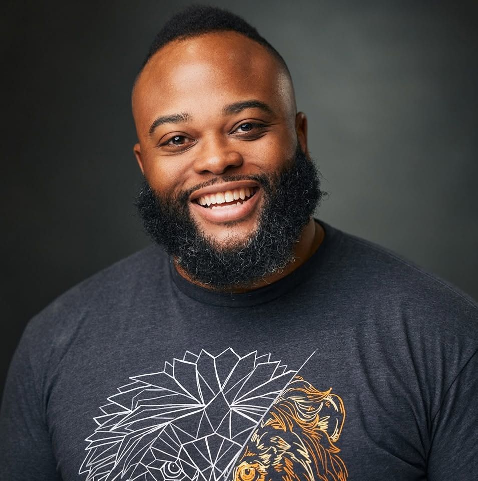 Smiling Black man with beard wearing gray graphic tee featuring lion design, professional headshot photo.