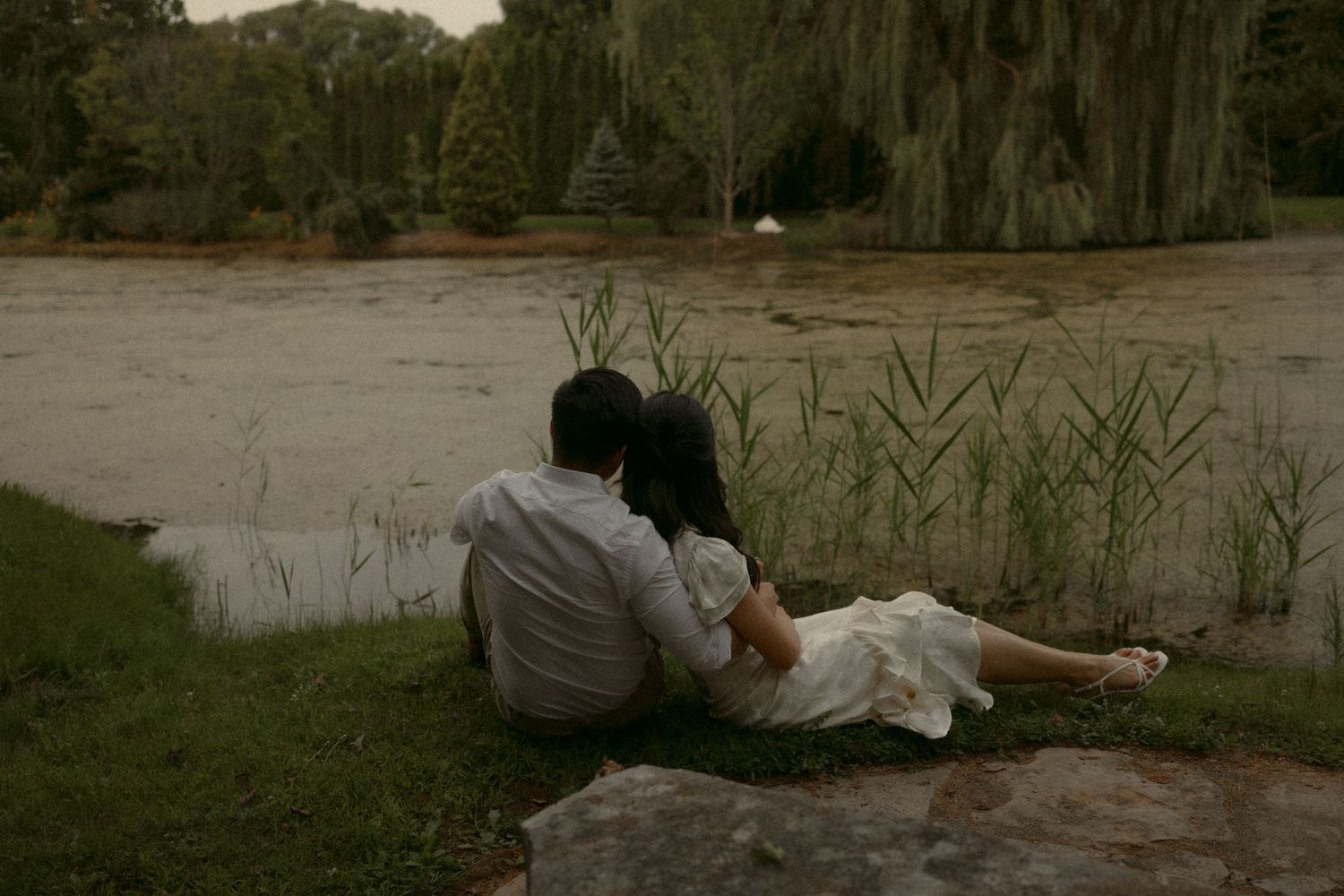 Couple sitting together on rocks by a tranquil pond surrounded by lush green trees in a romantic outdoor setting. Whistling Gardens Ontario
