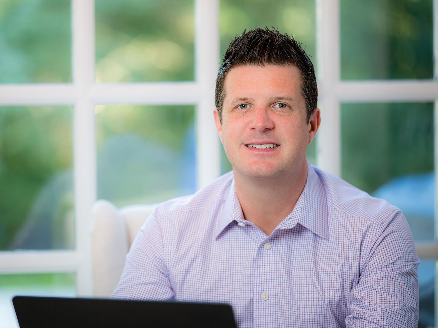 Professional man in light purple shirt smiling at desk with laptop in bright modern home office setting.