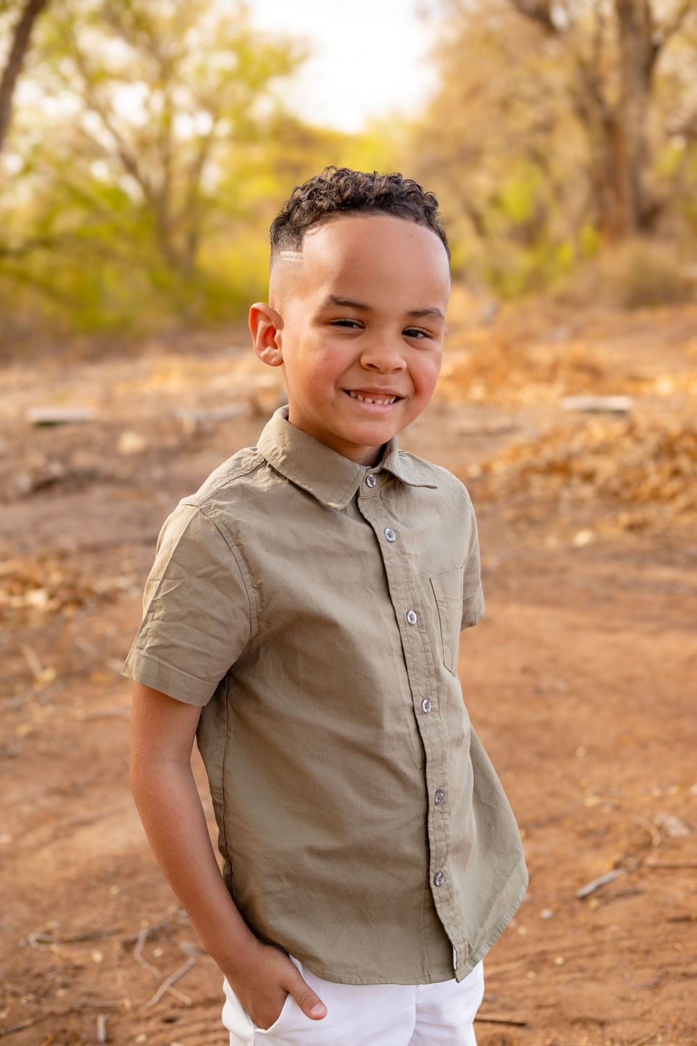 Smiling young boy in olive green shirt and white pants standing outdoors in a sunny natural setting.