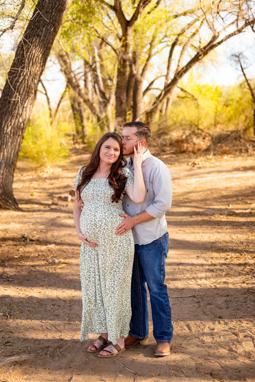 Couple poses outdoors during maternity photo shoot, man kissing woman's cheek as she cradles baby bump in floral dress.