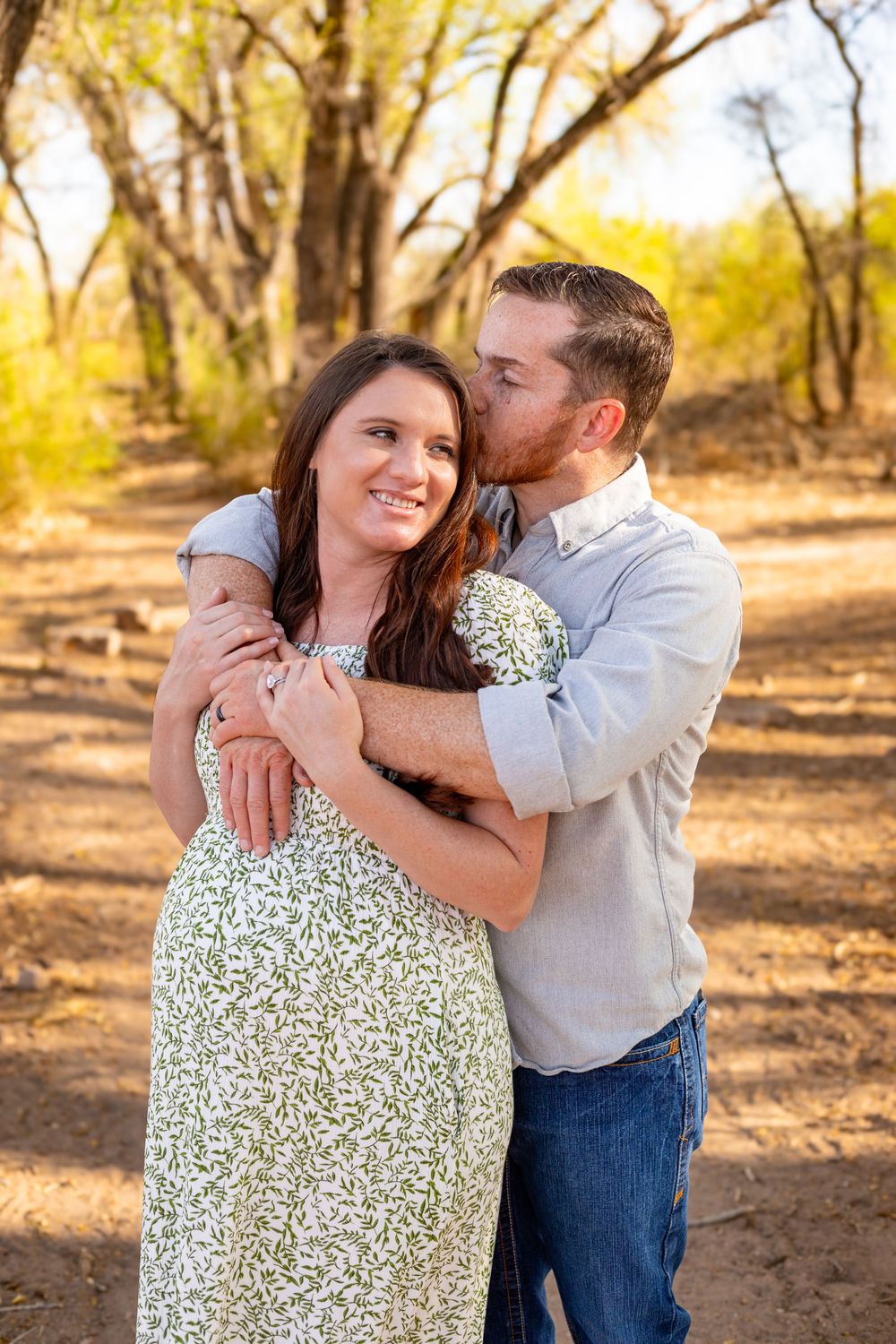 Couple embracing outdoors on a dirt path, woman in floral dress smiling as man kisses her cheek, surrounded by autumn trees.