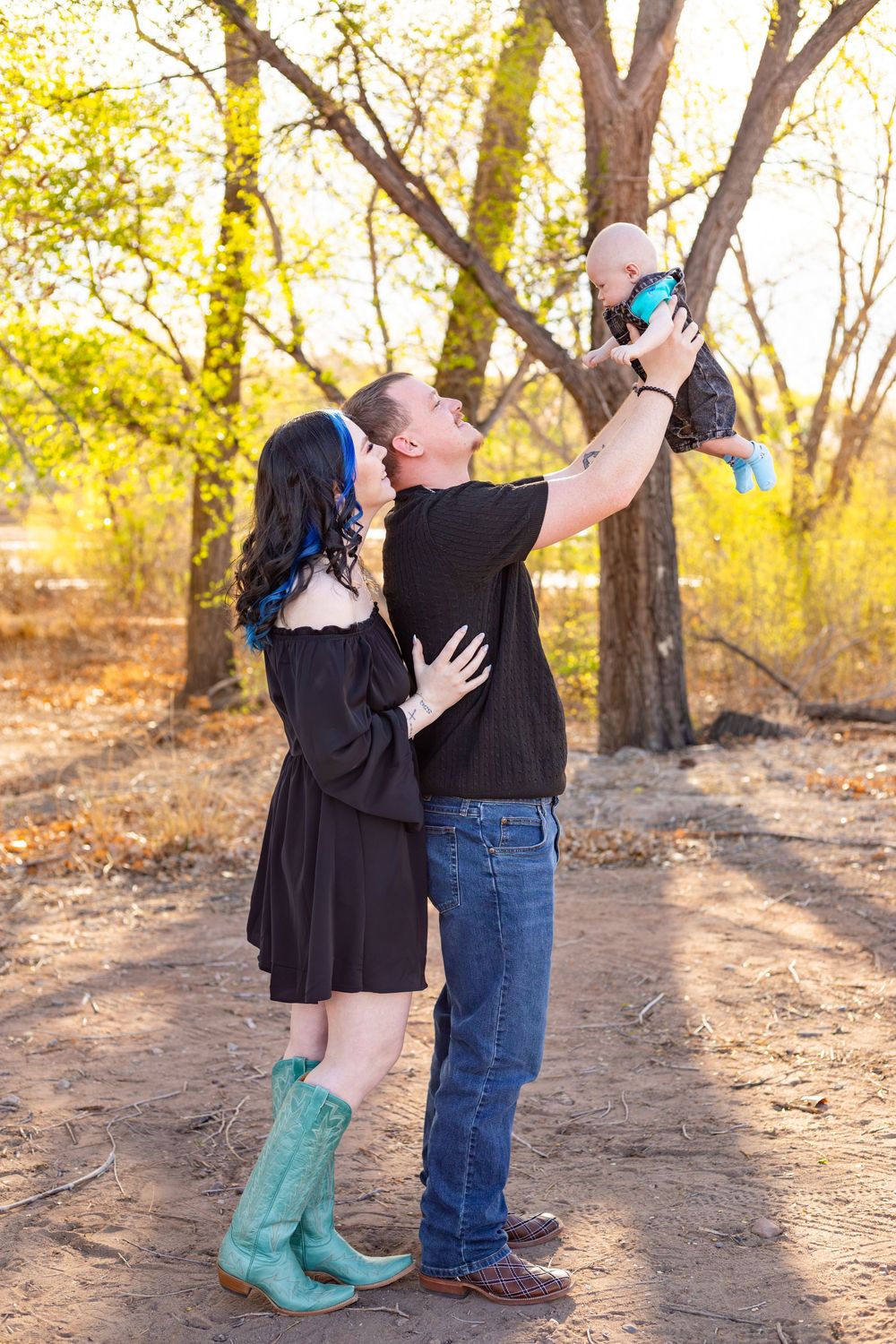 A couple kisses outdoors while lifting their baby in the air, surrounded by golden trees in a natural setting.