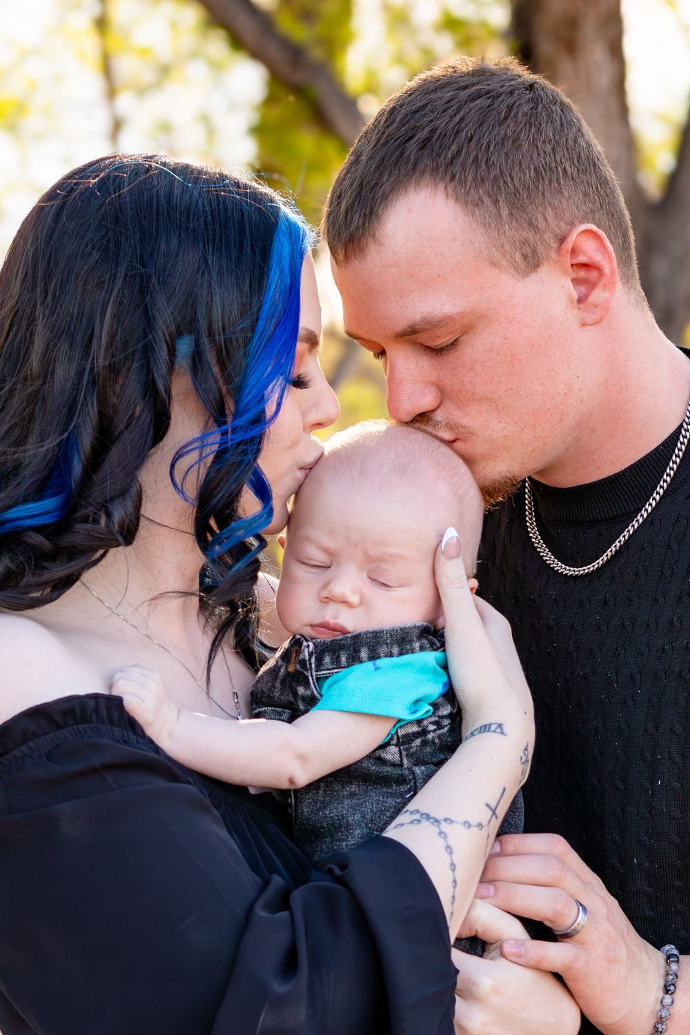 Parents with blue-streaked hair and chain necklace tenderly kiss their sleeping newborn baby outdoors.