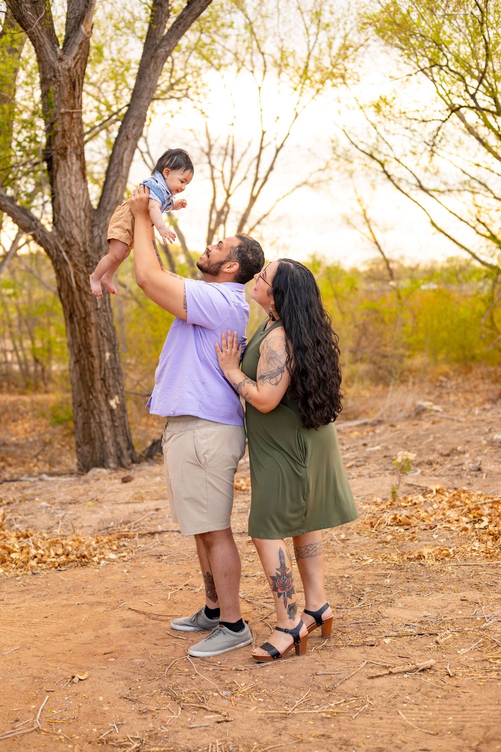 A couple lifts their baby outdoors among autumn trees, sharing a kiss in a warm, golden-lit natural setting.