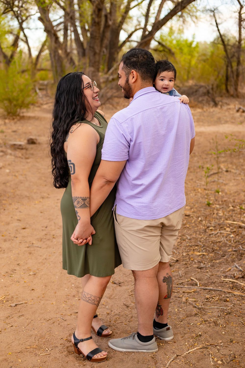 A young Hispanic family poses outdoors, mom in green dress, dad holding baby, surrounded by desert trees.