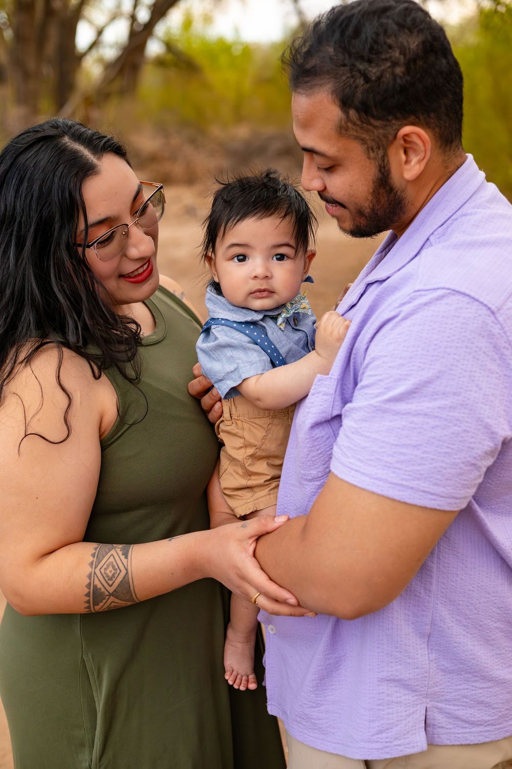 A young family with a baby boy poses outdoors, mom in green dress with tattoos, dad in lavender shirt.