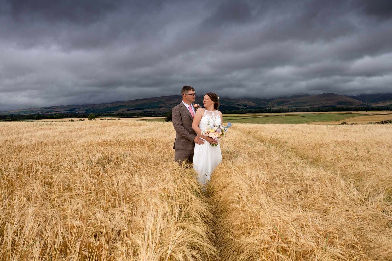 Bride and groom embracing in golden wheat field with dramatic stormy sky and rolling hills in background.