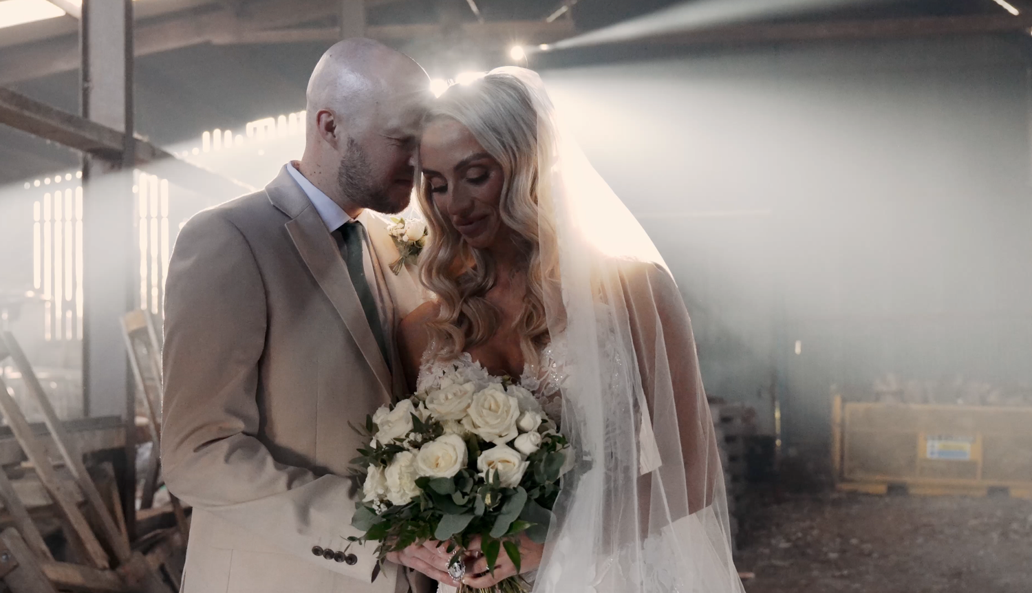Bride and groom share an intimate moment in a rustic venue, bride holding white roses bouquet with veil and soft light.
