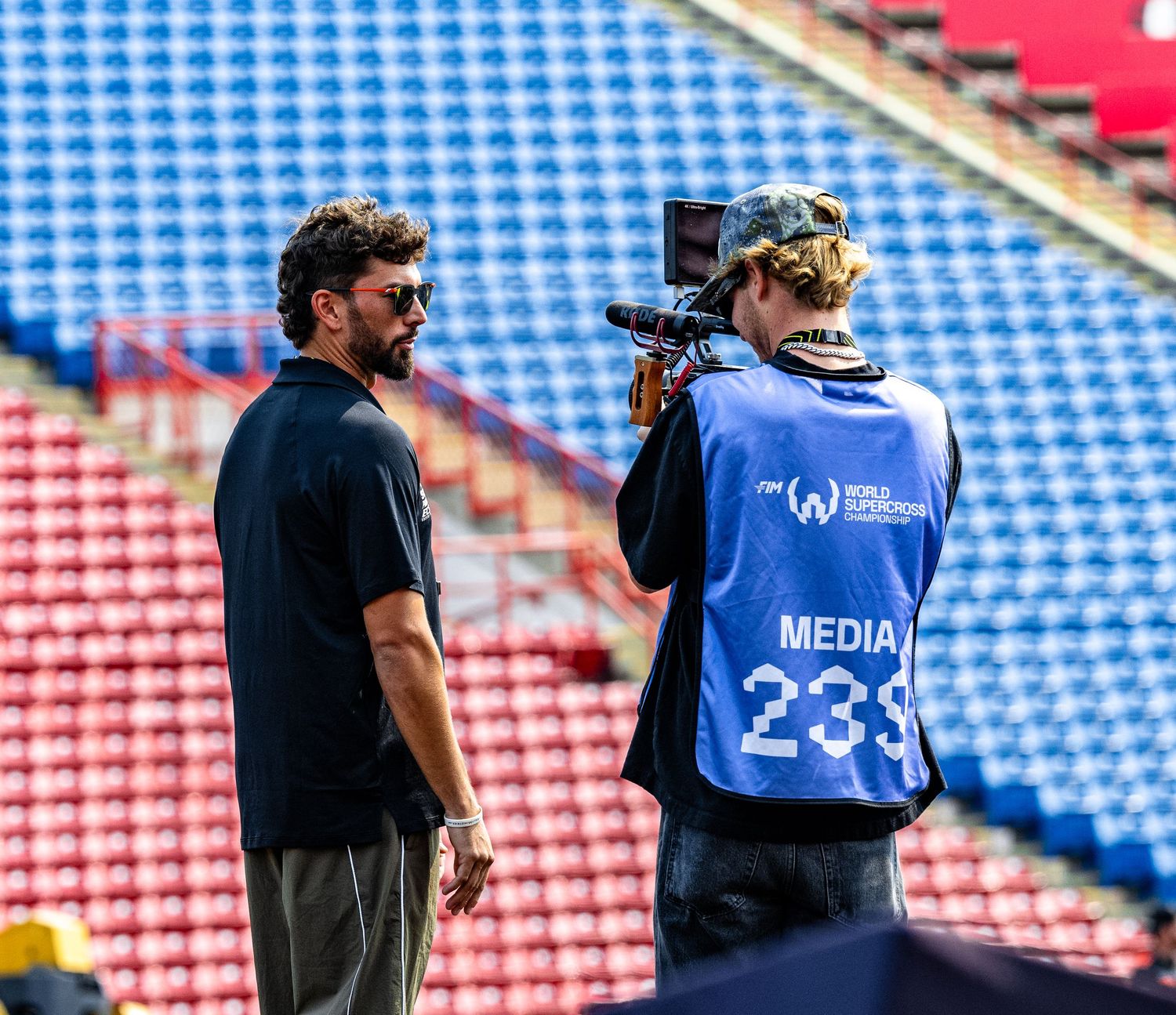 A media cameraman with accreditation badge films an interview with a man at a sports stadium.