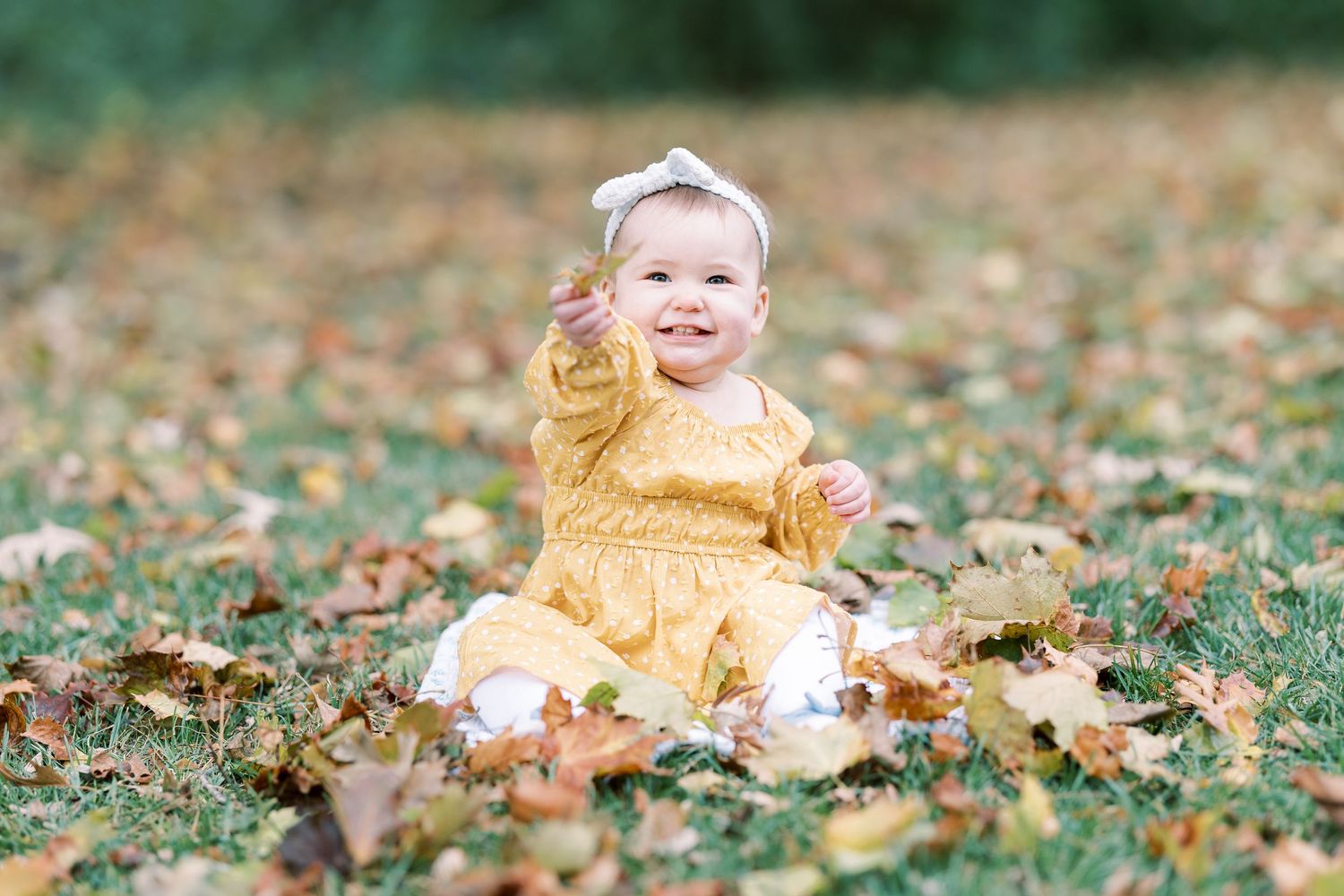 Smiling baby girl in yellow dress sitting on grass covered with autumn leaves outdoors.