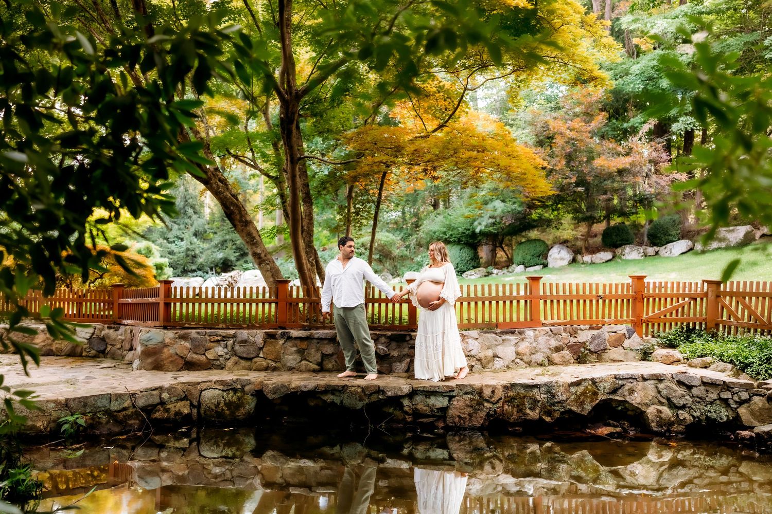 Couple stands on stone bridge over pond in lush garden with autumn foliage and wooden fence backdrop in Hamden, Connecticut
