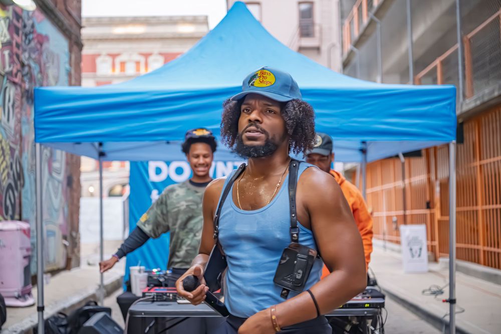 Man in blue tank top and cap works outdoor DJ setup under blue tent in urban alley setting.