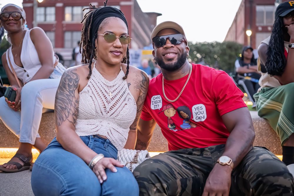 A smiling couple poses together outdoors at a festive evening event, woman with tattoos in white top and man in red shirt.