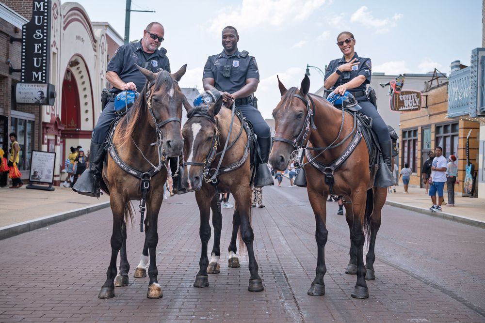 Three mounted police officers on brown horses patrol a busy street near a museum on a sunny day.