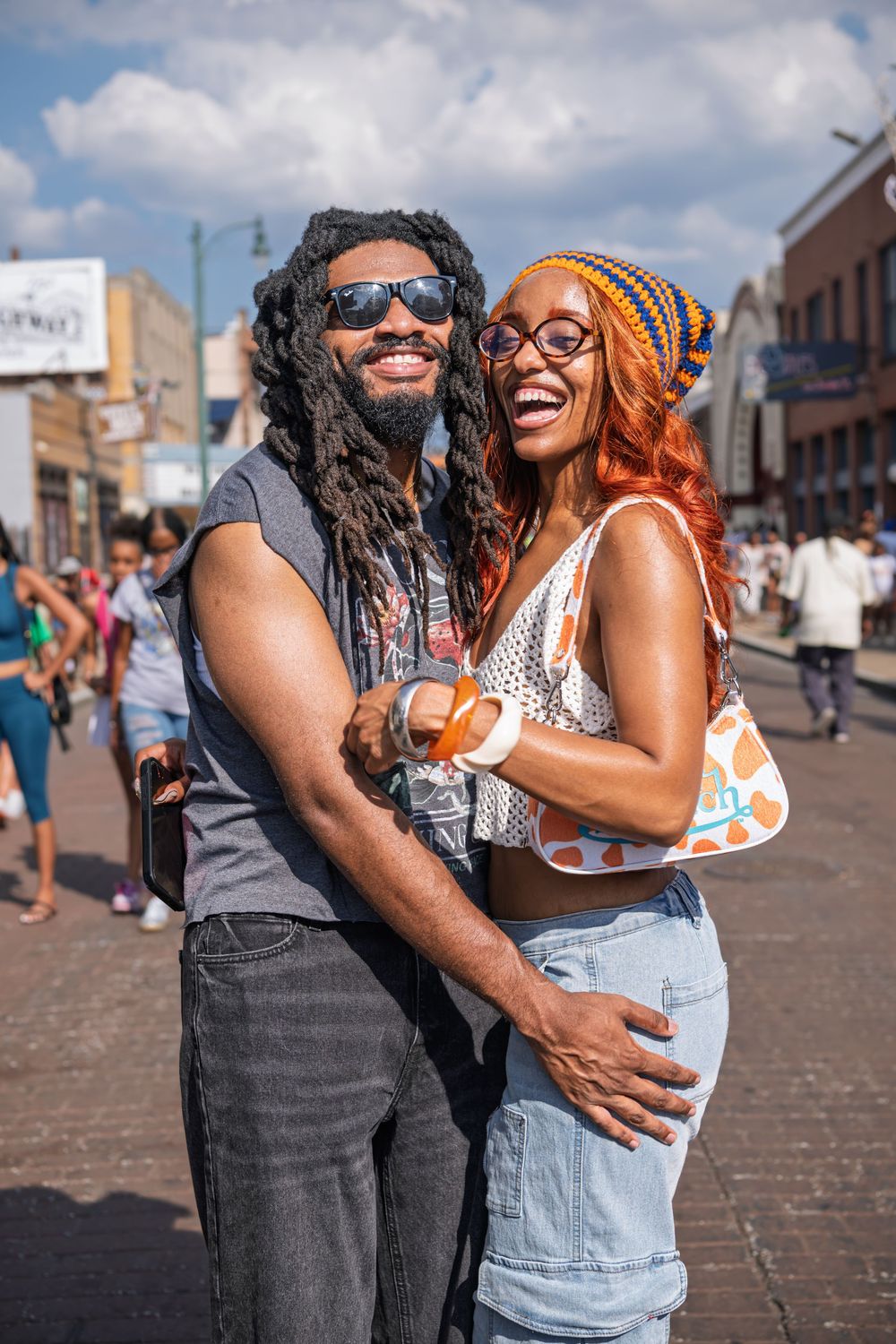A smiling couple with dreadlocks embraces on a sunny street, wearing colorful summer outfits and sunglasses.