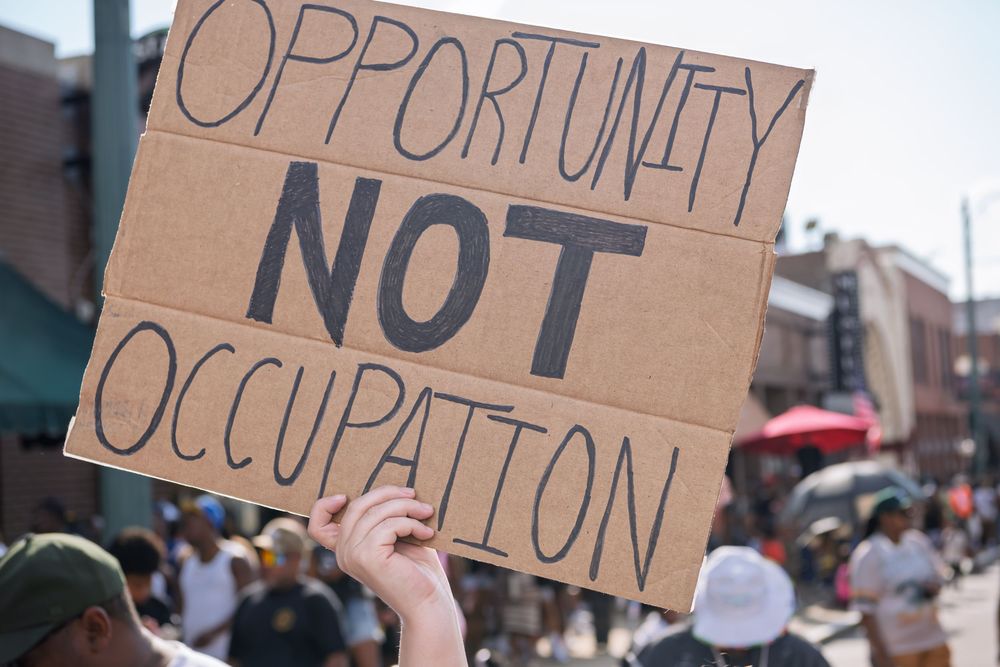 Protest sign reading 'Opportunity Not Occupation' held up at a street demonstration with crowd in background.
