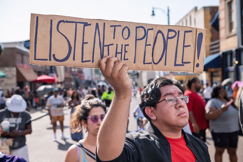 Protestor holds cardboard sign reading Listen To The People at outdoor demonstration on busy street.
