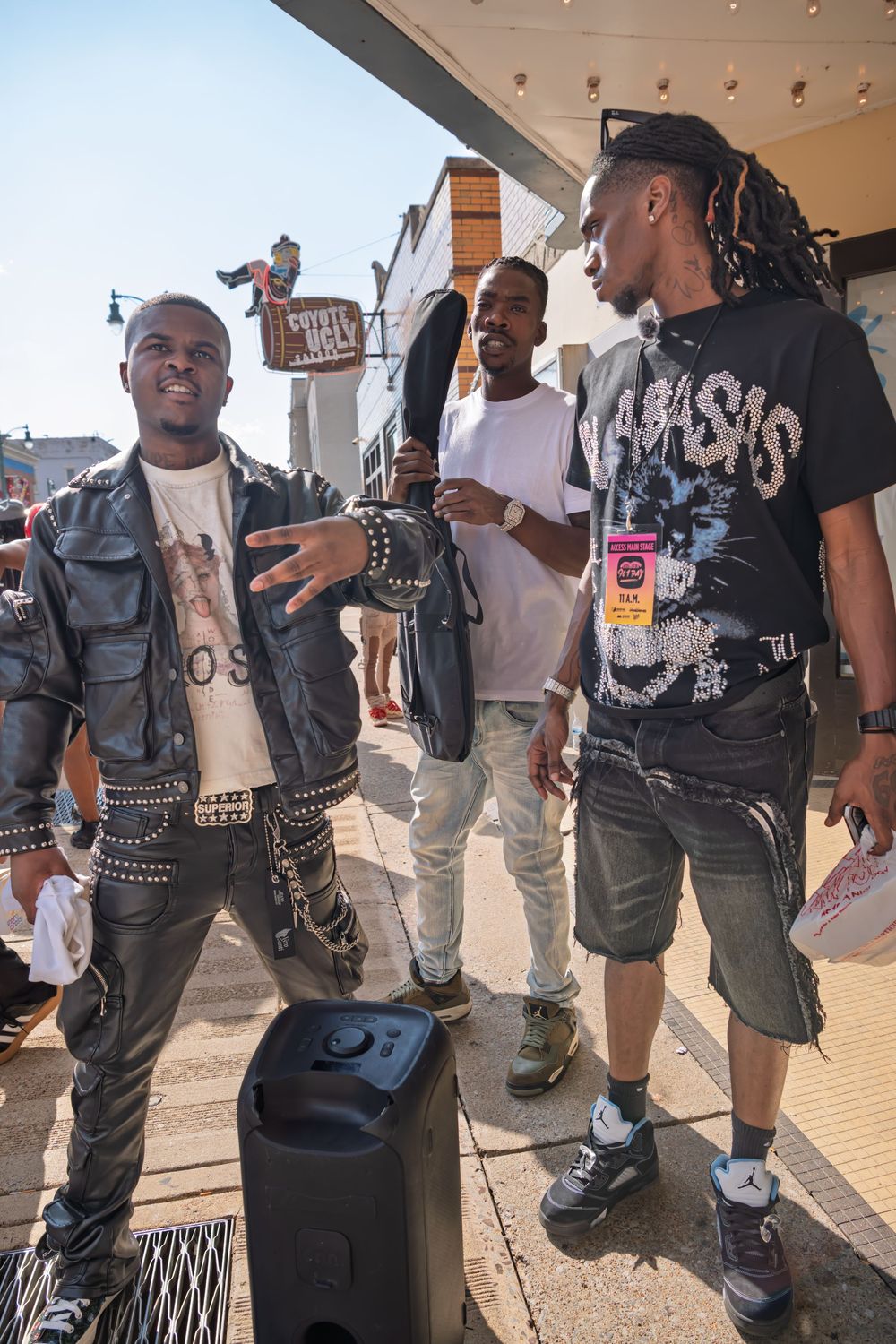 Three young men in streetwear socialize outside a venue on a sunny day, one wearing a studded leather jacket.