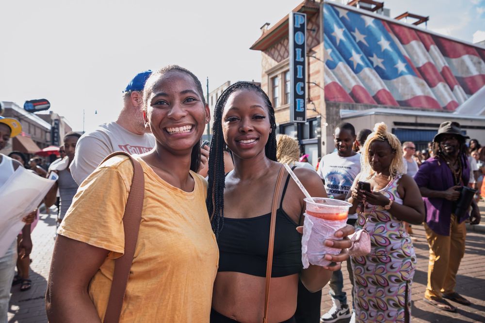 Two women smiling at an outdoor festival, one holding a pink drink, with an American flag mural in the background.