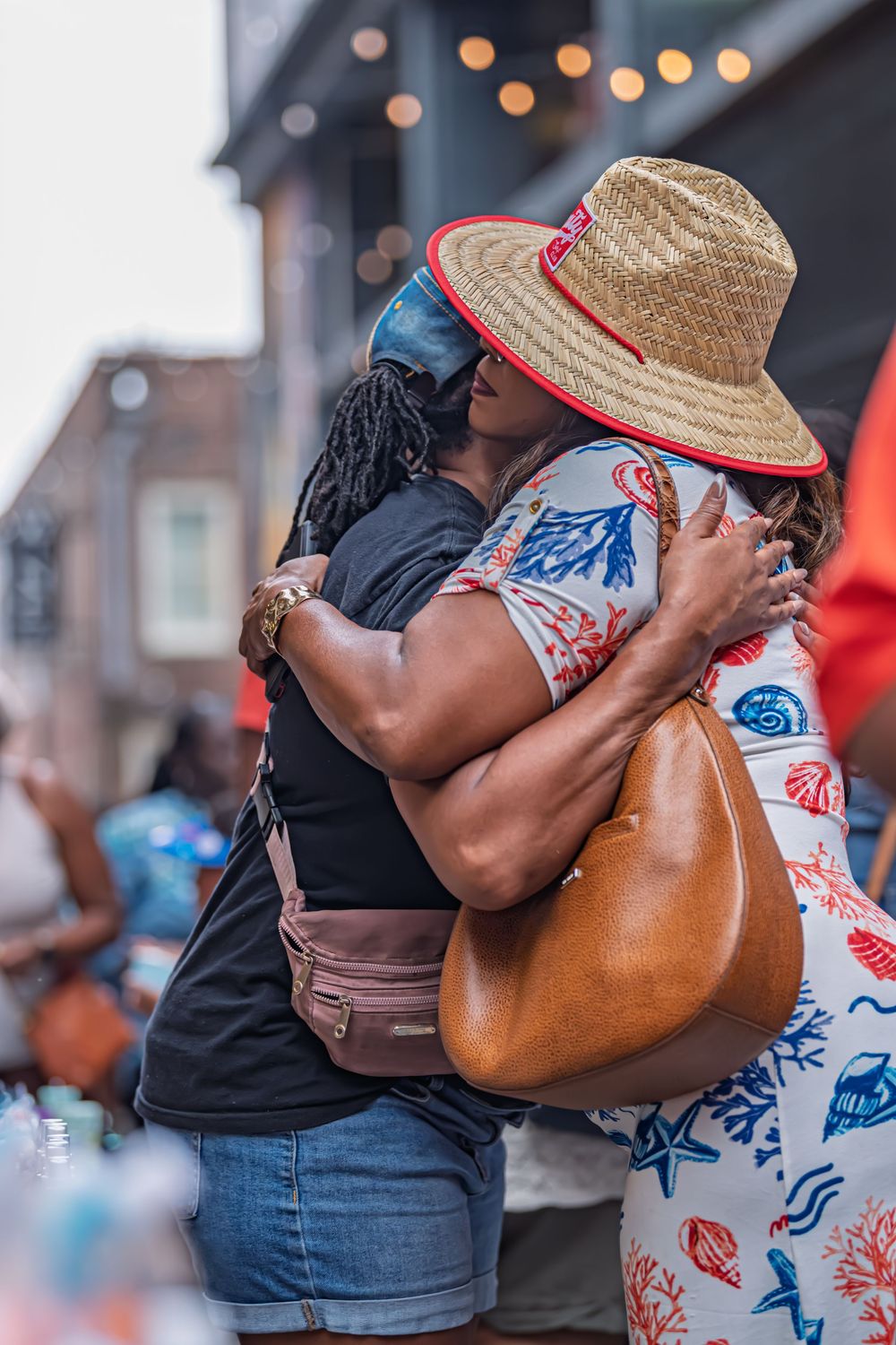 Two women share a warm embrace at an outdoor street festival, one wearing a straw sun hat and floral dress.