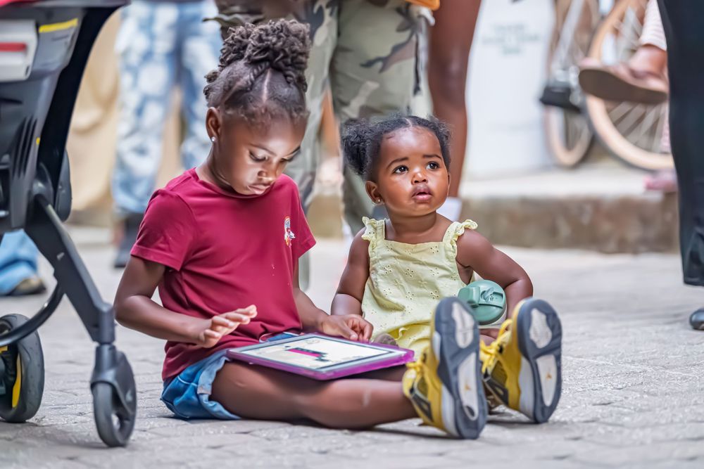 Two young girls sitting on pavement, one reading a colorful book outdoors near a stroller.