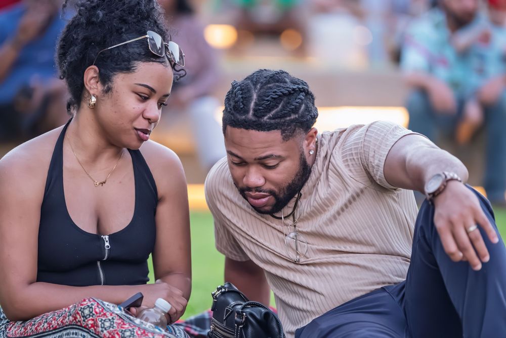 Two people sitting outdoors at an evening event, looking down together at something in a casual, intimate moment.